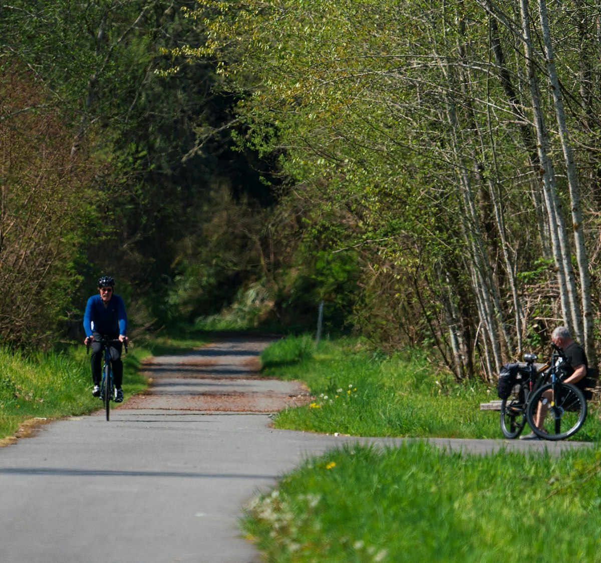 A biker rides along the pathway of the Yelm-Rainier-Tenino trail while another biker looks on from a bench.