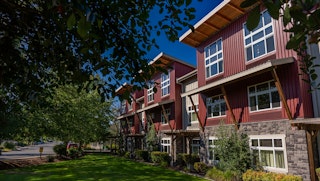 Modern red-sided hotel building with large windows, stone accents, and landscaped lawn under blue sky.